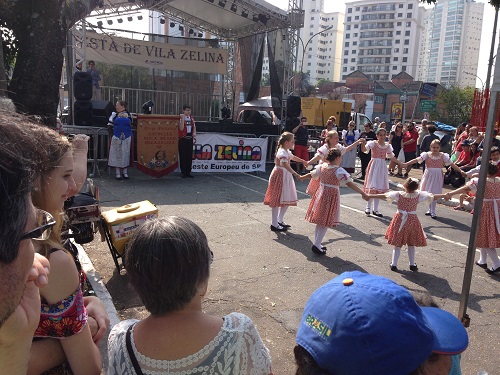 Imagem mostra o palco da festa de Vila Zelina ao fundo. Na frente, um grupo de dança faz uma apresentação.